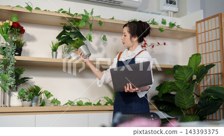 Asian female florist holding laptop while checking potted plants in store 143393375