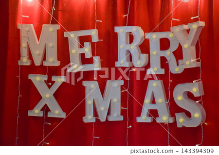 Lighted marquee letters spelling MERRY XMAS hang on a red curtain, with vertical string lights draped around them, creating a festive holiday decoration scene Lighted marquee letters spelling MERRY XMAS hang on a red curtain, with vertical string lights draped around them, creating a festive holiday decoration scene 143394309