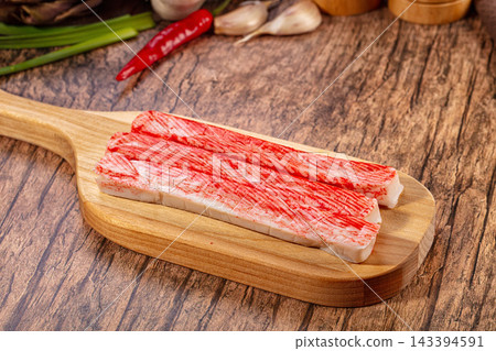 Crab stick seafood product on wooden cutting board with red and white striped texture, garlic and chili in background, kitchen preparation scene 143394591