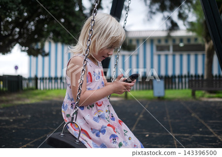 Little Girl Sitting on Swing and Looking at Smartphone Outdoors Little Girl Sitting on Swing and Looking at Smartphone Outdoors 143396500