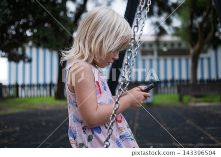 Little Girl Sitting on Swing and Looking at Smartphone Outdoors 143396504