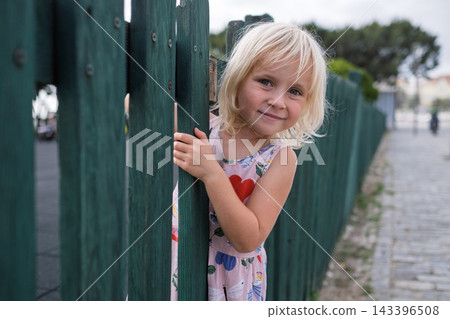 Cute Little Blonde Girl Peeking Behind Green Wooden Fence Outdoors Cute Little Blonde Girl Peeking Behind Green Wooden Fence Outdoors 143396508