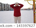Front view of a mature woman in red jacket warming up at snowy public sports ground in winter. 143397627