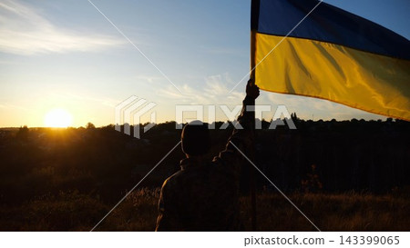 Male military in uniform lifting blue-yellow banner in honor of the victory against russian aggression at sunset. Young soldier of ukrainian army raised a waving flag of Ukraine at countryside Male military in uniform lifting blue-yellow banner in honor of the victory against russian aggression at sunset. Young soldier of ukrainian army raised a waving flag of Ukraine at countryside 143399065