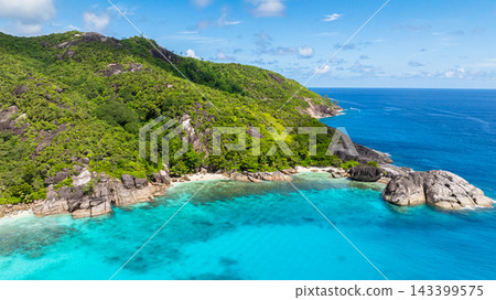Expansive green hills covered in dense vegetation meet the wide expanse of clear blue ocean with rocky formations. Seychelles, Mahe. Expansive green hills covered in dense vegetation meet the wide expanse of clear blue ocean with rocky formations. Seychelles, Mahe. 143399575