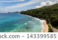 Waves gently lap on a sandy shore surrounded by green hills and clear blue waters, under a bright sky. Seychelles, Mahe. Anse Takamaka Beach. 143399585