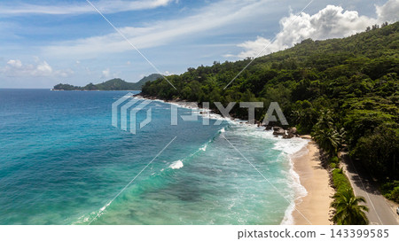 Waves gently lap on a sandy shore surrounded by green hills and clear blue waters, under a bright sky. Seychelles, Mahe. Anse Takamaka Beach. Waves gently lap on a sandy shore surrounded by green hills and clear blue waters, under a bright sky. Seychelles, Mahe. Anse Takamaka Beach. 143399585