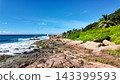 Rocky coastline with turquoise waves crashing against the shore, surrounded by lush green vegetation under a blue sky. Seychelles, La Digue. 143399593