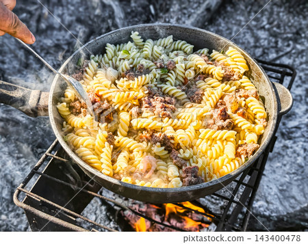 Pasta is being prepared with meat on a campfire at a nature site. Steam rises from the pot as food is mixed. This is a group activity enjoyed in the 143400478