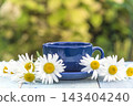 Blue cup of tea and bouquet of white daisy (chamomile) flowers on a wooden table against blurred natural background 143404240