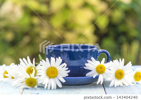 Blue cup of tea and bouquet of white daisy (chamomile) flowers on a wooden table against blurred natural background 143404240
