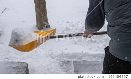 Snow shoveling in winter landscape, individual using yellow shovel to clear snow from walkway, showcasing winter maintenance and outdoor activity Snow shoveling in winter landscape, individual using yellow shovel to clear snow from walkway, showcasing winter maintenance and outdoor activity 143404367