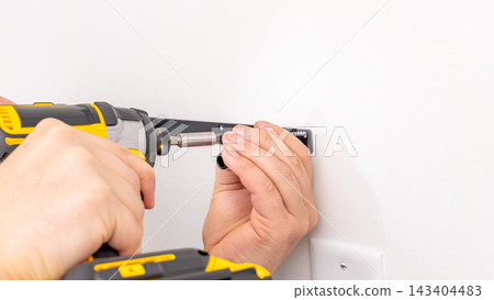 Mans hands tightening screw on black metal shelf bracket attached to white wall with cordless drill. Home improvement, final tightening, DIY shelf installation completion, secure fixture. 143404483