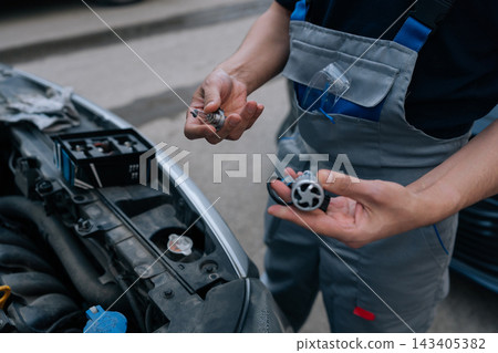 Auto mechanic comparing traditional halogen bulb with modern led headlight bulb in hand, standing near car engine bay, preparing for vehicle lighting upgrade or repair. Auto mechanic comparing traditional halogen bulb with modern led headlight bulb in hand, standing near car engine bay, preparing for vehicle lighting upgrade or repair. 143405382
