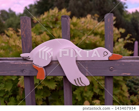 Pastel unicorn toy leaning on weathered wooden fence outdoors at dusk with soft pink sky, green bushes, and warm light in calm suburban garden 143407934