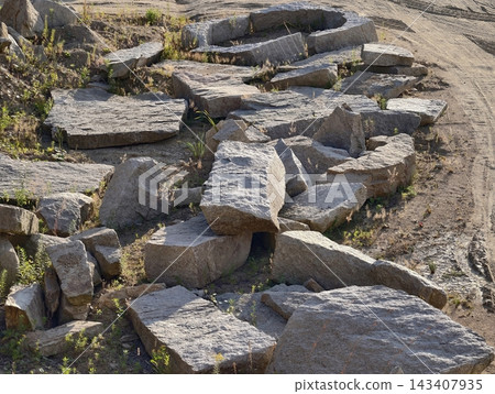 Cracked granite rubble and weathered concrete blocks scattered along sunlit dirt shoulder with tufts grasses and hardy weeds growing in crevices under warm afternoon light 143407935