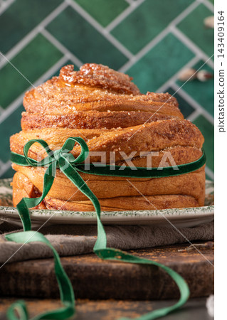 A vertical shot of a flaky puff pastry Easter bread coated in cinnamon sugar and tied with a green ribbon against a stylish emerald tiled background A vertical shot of a flaky puff pastry Easter bread coated in cinnamon sugar and tied with a green ribbon against a stylish emerald tiled background 143409164