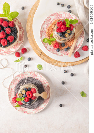 A bright and airy high-angle flat lay featuring two jars of layered overnight oats with chia seeds and granola, topped with fresh raspberries and blueberries. The scene is artfully arranged on a A bright and airy high-angle flat lay featuring two jars of layered overnight oats with chia seeds and granola, topped with fresh raspberries and blueberries. The scene is artfully arranged on a 143409165