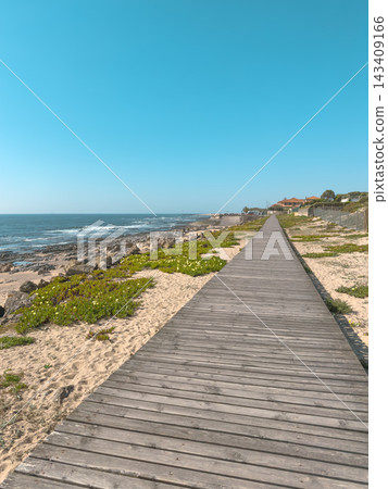A long rustic wooden boardwalk stretches along the rocky shoreline and sandy dunes of Praia da Granja in Porto Portugal under a clear blue sky during a sunny day A long rustic wooden boardwalk stretches along the rocky shoreline and sandy dunes of Praia da Granja in Porto Portugal under a clear blue sky during a sunny day 143409166
