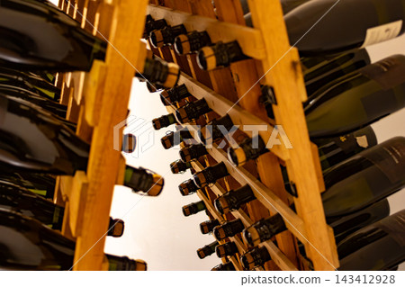 Sparkling wine bottles aging in wooden riddling racks, undergoing the traditional method of fermentation in a cellar, developing complex flavors for future enjoyment Sparkling wine bottles aging in wooden riddling racks, undergoing the traditional method of fermentation in a cellar, developing complex flavors for future enjoyment 143412928