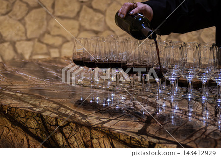 Hand pouring red wine from a bottle into tasting glasses on a beautiful marble counter, preparing for an exclusive winery event or vintage barrel aging sampling Hand pouring red wine from a bottle into tasting glasses on a beautiful marble counter, preparing for an exclusive winery event or vintage barrel aging sampling 143412929