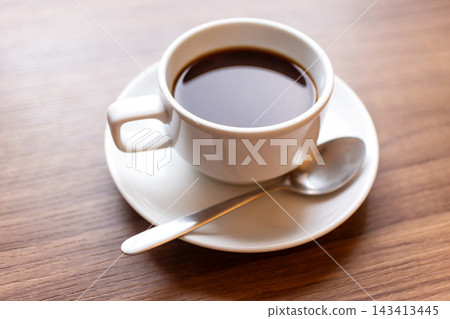 Fresh black coffee cup with spoon on saucer over wooden table in morning sunlight Fresh black coffee cup with spoon on saucer over wooden table in morning sunlight 143413445