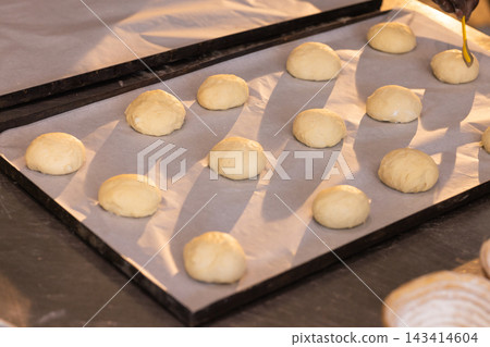 Parchment pan holding raw rolls at bakery counter with yellow brush and floured proofing basket Parchment pan holding raw rolls at bakery counter with yellow brush and floured proofing basket 143414604
