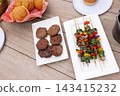White plates sitting on picnic table holding six burger patties, vegetable skewers and bread basket 143415232