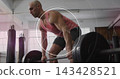 Lifting bald man gripping loaded barbell at gym in coral tank and dark shorts, circular overlay 143428521