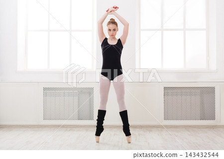 A ballet dancer stands in a studio with large windows. She stretches her arms above her head while wearing a black leotard and tights. The bright light enters through the windows. 143432544