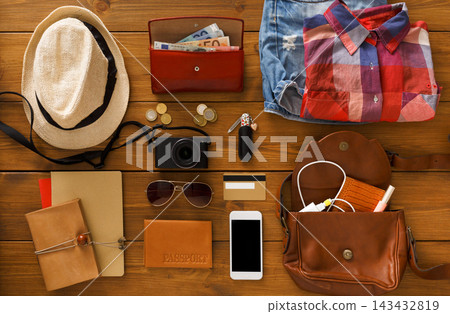 A collection of items for a day trip is laid out on a wooden table. It includes a camera, clothes, money, coins, sunglasses, and a smartphone beside a brown bag and a hat. A collection of items for a day trip is laid out on a wooden table. It includes a camera, clothes, money, coins, sunglasses, and a smartphone beside a brown bag and a hat. 143432819
