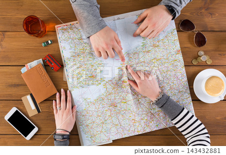 Two people are looking at a map on a wooden table while planning a trip. Drinks are nearby along with a phone, a wallet, and some coins. The atmosphere is relaxed and focused. Two people are looking at a map on a wooden table while planning a trip. Drinks are nearby along with a phone, a wallet, and some coins. The atmosphere is relaxed and focused. 143432881