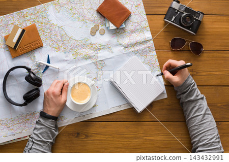 A person sits at a table with a map spread out, writing in a notebook. There is a cup of coffee, headphones, a camera, coins, and a passport nearby. A person sits at a table with a map spread out, writing in a notebook. There is a cup of coffee, headphones, a camera, coins, and a passport nearby. 143432991