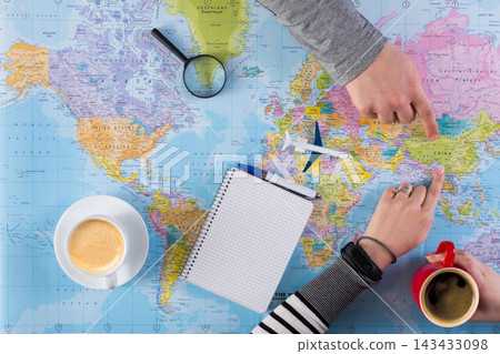 Two people are pointing at locations on a world map spread on a table. A notebook and pens are present along with two cups of coffee. The setting suggests planning a journey. 143433098