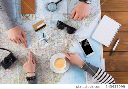 A group of friends points at locations on a map spread out on a wooden table. A phone, notebook, and a coffee cup are on the table. Money and a camera are visible nearby. A group of friends points at locations on a map spread out on a wooden table. A phone, notebook, and a coffee cup are on the table. Money and a camera are visible nearby. 143433099