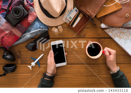 Hands hold a phone and coffee cup above a table with a camera, sunglasses, currency, map, and travel items. This scene shows someone getting ready for a trip. Hands hold a phone and coffee cup above a table with a camera, sunglasses, currency, map, and travel items. This scene shows someone getting ready for a trip. 143433118