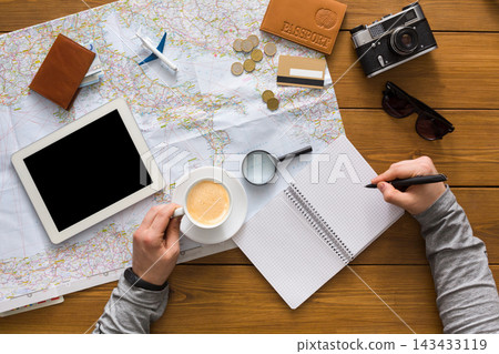 A person is holding a cup of coffee while making notes on a notepad. Surrounding are a map, a tablet, a camera, and travel items. Coins and a passport are visible on the table. A person is holding a cup of coffee while making notes on a notepad. Surrounding are a map, a tablet, a camera, and travel items. Coins and a passport are visible on the table. 143433119