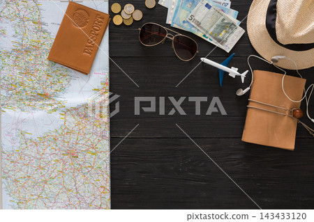 A map is spread out on a wooden table with a passport, coins, currency, sunglasses, and a model airplane. Travel items are placed beside a notebook and earphones, indicating trip preparations. A map is spread out on a wooden table with a passport, coins, currency, sunglasses, and a model airplane. Travel items are placed beside a notebook and earphones, indicating trip preparations. 143433120