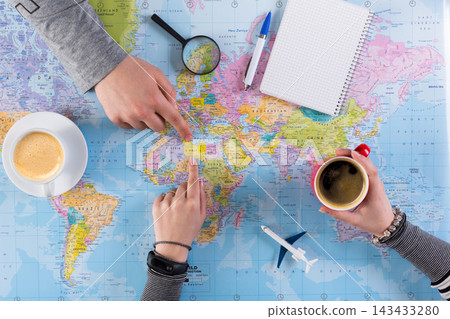 Two people are discussing travel plans at a table. They are pointing at different locations on a world map. Coffee cups are on the table along with a notebook and pens. Two people are discussing travel plans at a table. They are pointing at different locations on a world map. Coffee cups are on the table along with a notebook and pens. 143433280