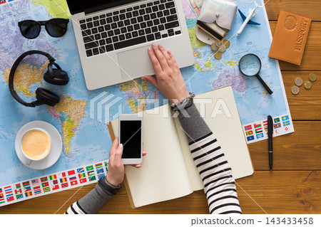 A person uses a laptop and holds a smartphone while sitting at a wooden table. A world map and travel items are present. Coffee sits nearby. The scene shows travel planning and preparation. A person uses a laptop and holds a smartphone while sitting at a wooden table. A world map and travel items are present. Coffee sits nearby. The scene shows travel planning and preparation. 143433458