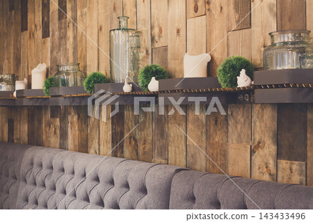 Shelves with jars, plants, and small decorative items are arranged against a wooden wall. A comfortable grey tufted bench is placed below. This is a warm and inviting spot in a cafe. 143433496