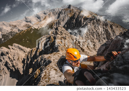 Close-up of a focused solo male climber ascending a rugged rock wall on a via ferrata in the Austrian Alps near Innsbruck, using safety equipment and climbing gear. Close-up of a focused solo male climber ascending a rugged rock wall on a via ferrata in the Austrian Alps near Innsbruck, using safety equipment and climbing gear. 143433523