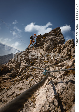 Solo male climber ascending a rugged alpine via ferrata route in the Austrian Alps near Innsbruck, Tyrol, with a steel safety cable leading through dramatic rocky terrain. Solo male climber ascending a rugged alpine via ferrata route in the Austrian Alps near Innsbruck, Tyrol, with a steel safety cable leading through dramatic rocky terrain. 143433524
