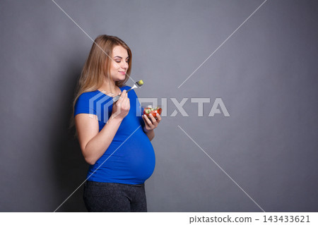 A woman who is expecting is standing in a neutral studio space. She is smiling as she eats salad with a fork. The setting has a simple gray background and the woman is dressed in a blue top. A woman who is expecting is standing in a neutral studio space. She is smiling as she eats salad with a fork. The setting has a simple gray background and the woman is dressed in a blue top. 143433621