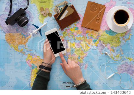A person is checking a smartphone while sitting at a table with a world map. Nearby, there is a cup of coffee, a wallet with cash and coins, and a passport. Travel gear is visible. A person is checking a smartphone while sitting at a table with a world map. Nearby, there is a cup of coffee, a wallet with cash and coins, and a passport. Travel gear is visible. 143433643