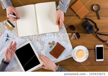 Two hands are seen working on travel plans using a map, tablet, notebook, and camera on a wooden table. A cup of coffee and sunglasses are also present, creating a lively setup for exploration. Two hands are seen working on travel plans using a map, tablet, notebook, and camera on a wooden table. A cup of coffee and sunglasses are also present, creating a lively setup for exploration. 143433672