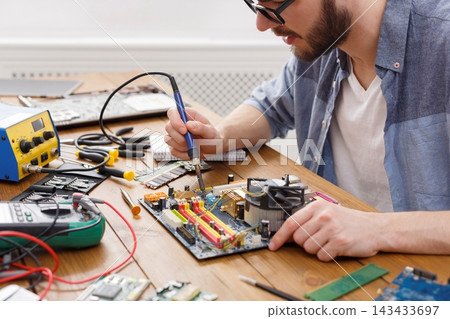 A technician repairs a computer motherboard at a workbench filled with tools and electronic components. He uses a soldering iron and examines the circuit connections closely. 143433697