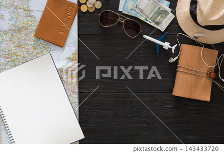 A map lays on a wooden table with a passport, money, sunglasses, a small airplane, and a notebook. Travel items are arranged for planning a trip. Accessories are present for convenience. A map lays on a wooden table with a passport, money, sunglasses, a small airplane, and a notebook. Travel items are arranged for planning a trip. Accessories are present for convenience. 143433720