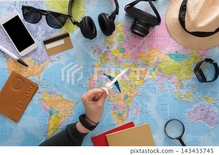 A hand holds a small airplane over a world map on a table filled with travel items. Nearby are a camera, headphones, smartphone, passport, and notebooks. Travel planning is in progress. A hand holds a small airplane over a world map on a table filled with travel items. Nearby are a camera, headphones, smartphone, passport, and notebooks. Travel planning is in progress. 143433745
