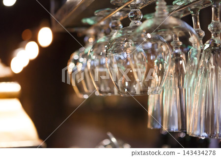 Crystal-clear glasses hang upside down from a bar shelf, illuminated by soft, warm lighting in a cozy setting. The atmosphere is inviting and relaxed, perfect for socializing. Crystal-clear glasses hang upside down from a bar shelf, illuminated by soft, warm lighting in a cozy setting. The atmosphere is inviting and relaxed, perfect for socializing. 143434278